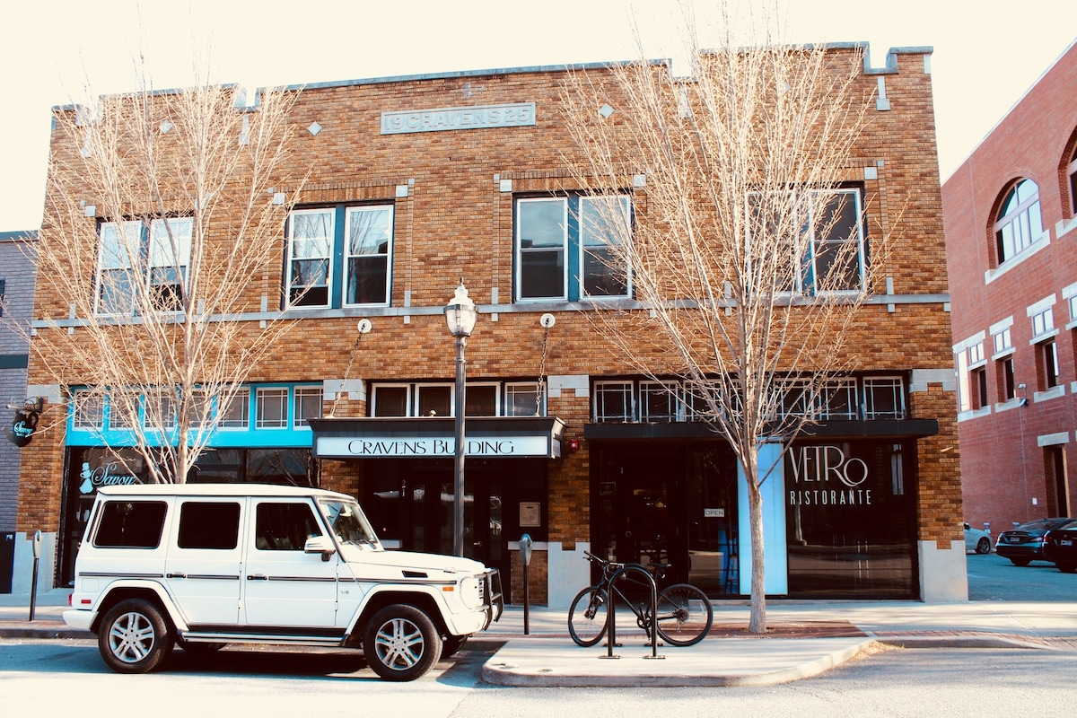 The exterior of the Craven's Building is showcased, featuring a brick façade with large windows and a central entrance. A white vehicle is parked nearby, alongside a bicycle. Leafless trees line the walkway, contributing to the downtown atmosphere.