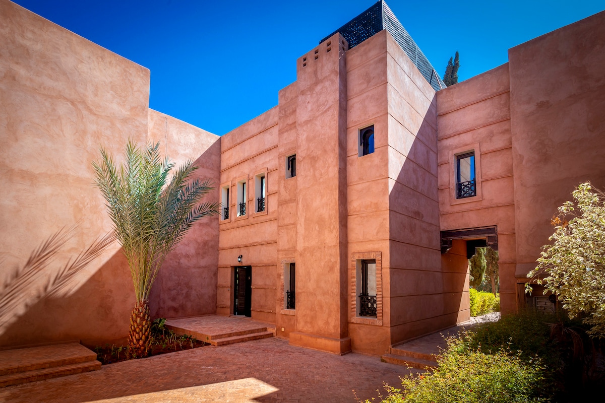 The exterior of the Riad features earthy-toned walls, showcasing traditional Moroccan architecture. A tall palm tree adds a touch of greenery, while light filters through the windows. The entrance is framed by stone pathways, leading into the tranquil courtyard.