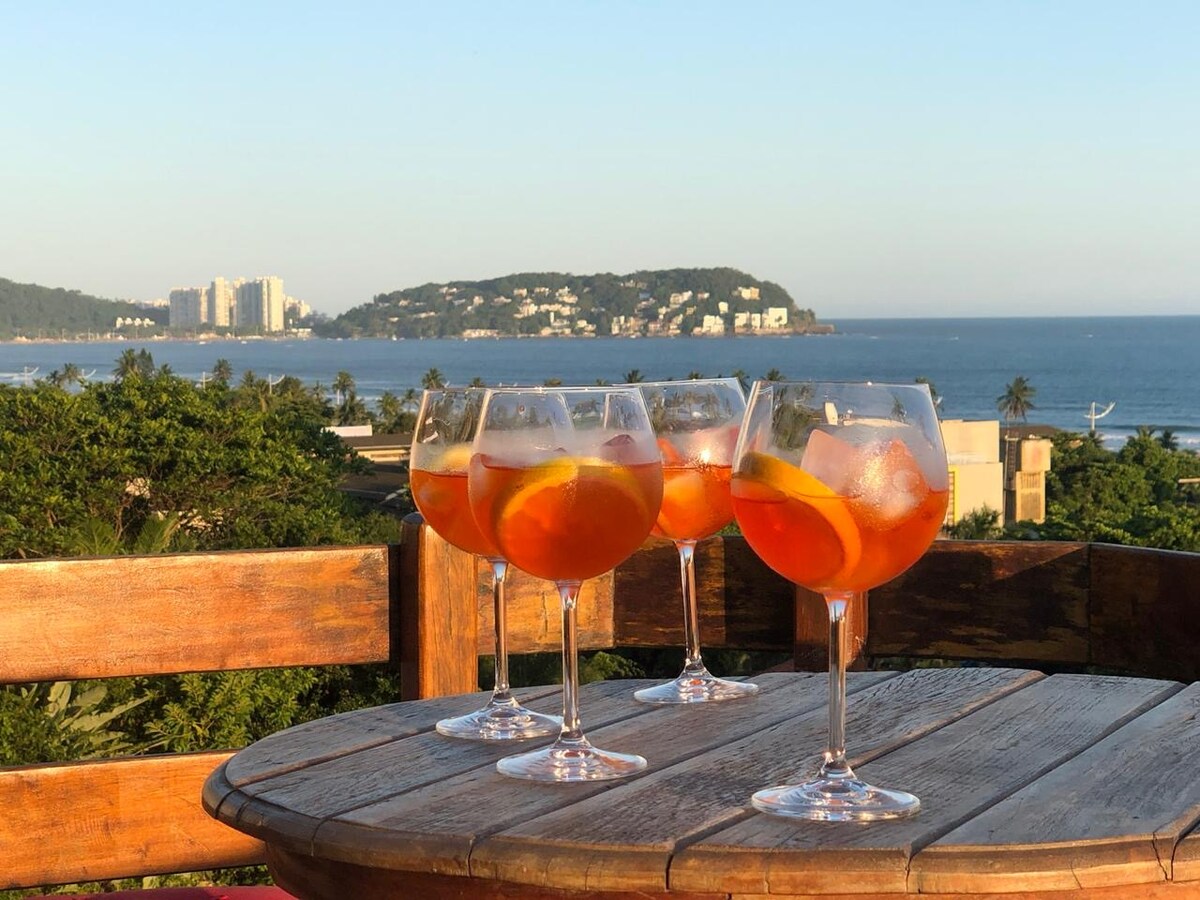 A scenic view of a coastline is captured, featuring glasses of cocktails resting on a wooden table. The drinks are garnished with orange slices, set against a vibrant sunset sky and the distant silhouette of hills and buildings along the shoreline.
