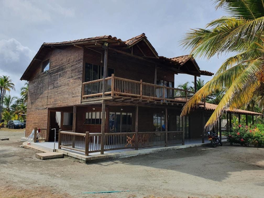 A two-story wooden house features a spacious deck with multiple seating areas. Large windows allow natural light to stream in, while a palm tree can be seen nearby. The structure is surrounded by sandy terrain and lush greenery, creating a cozy outdoor atmosphere.