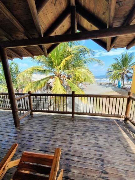 A shaded wooden balcony features a view of swaying palm trees and the ocean in the distance. Natural light floods the space, highlighting the wooden flooring and railings that provide an inviting outdoor area.