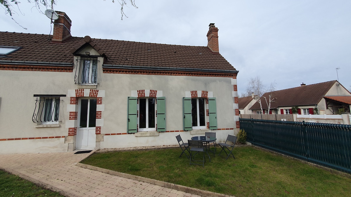 A charming exterior of a longère features light-colored walls with green shutters and a brick trim. A patio area with a small table and chairs is visible on the well-maintained lawn, providing a space for relaxation. A pathway leads to the entrance, enhancing accessibility.