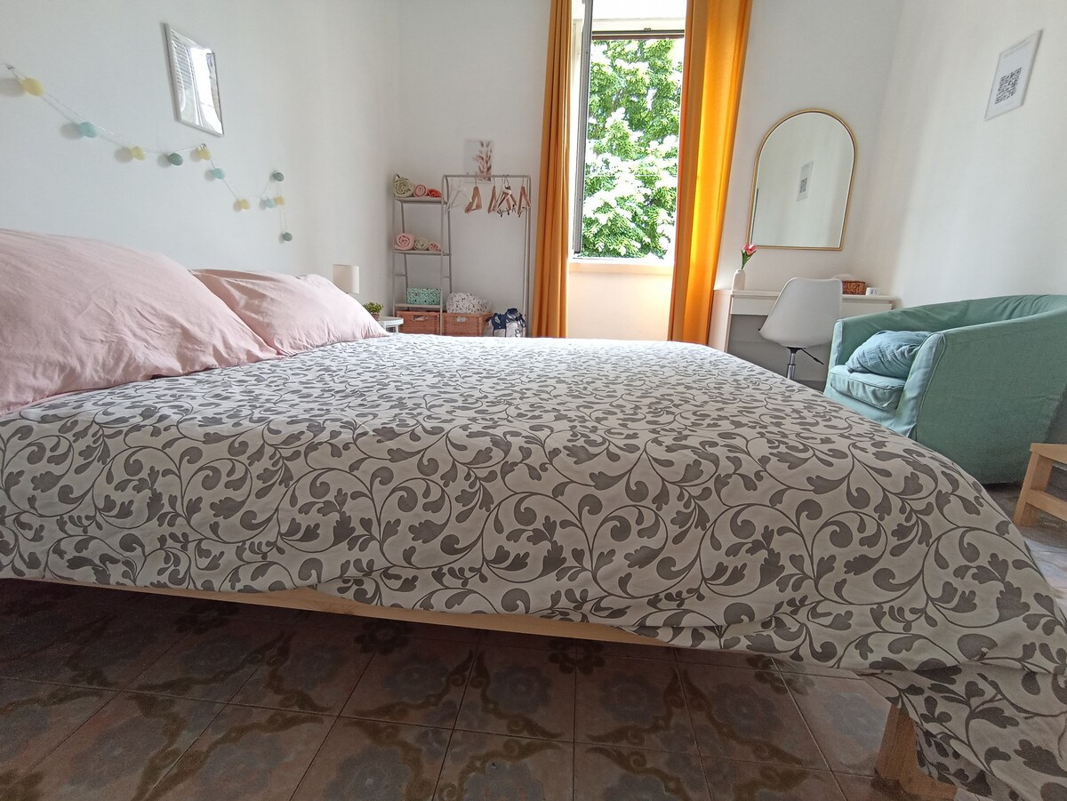 A spacious bedroom features a large bed with a patterned blanket and soft pink pillows. Natural light floods the room through a window dressed with light curtains. A cozy seating area is visible, along with a mirror and decorative elements adorning the walls.