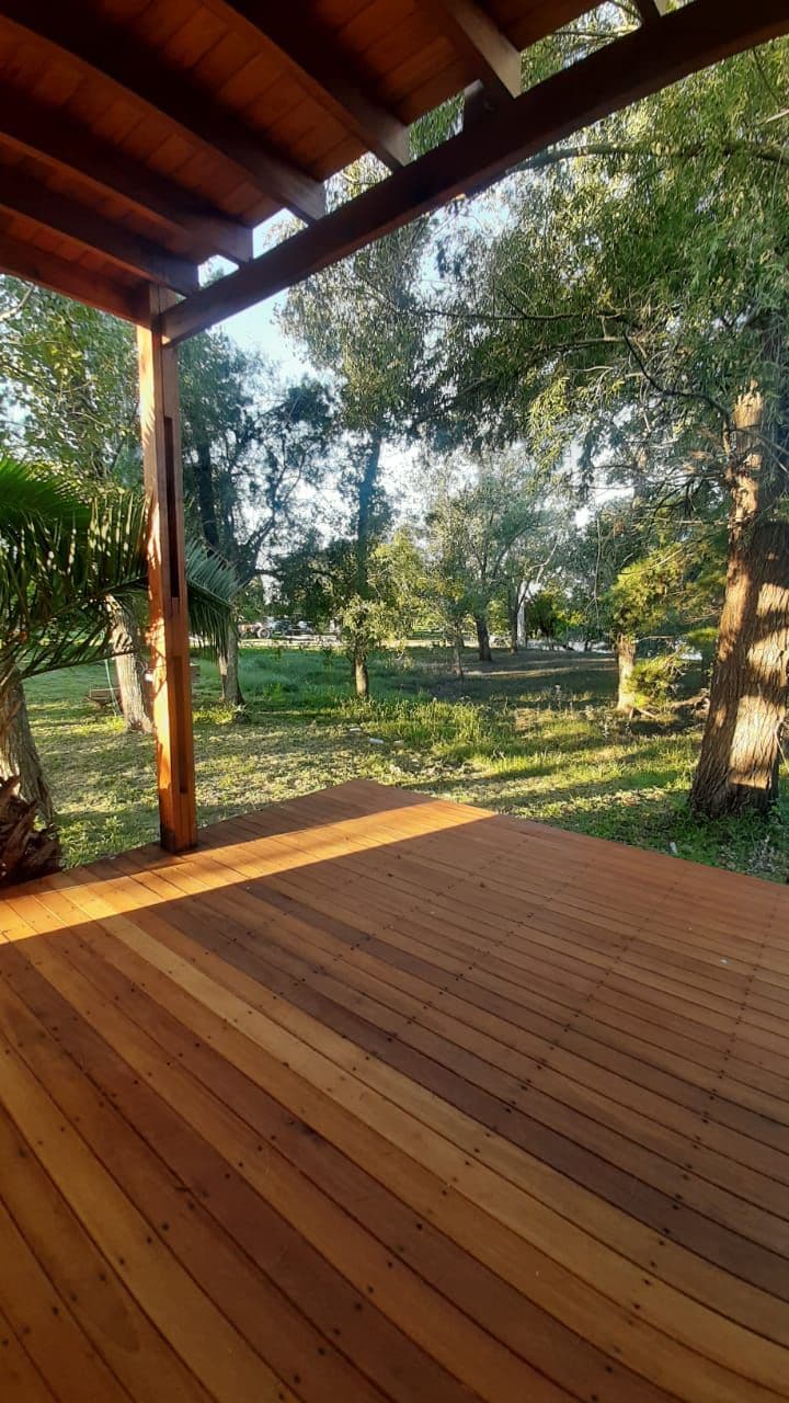 A wooden deck is shown surrounded by lush greenery and tall trees. Sunlight filters through the foliage, casting gentle shadows on the ground. The serene surroundings include an open area with grass and flowering plants, creating a peaceful outdoor space near the river.