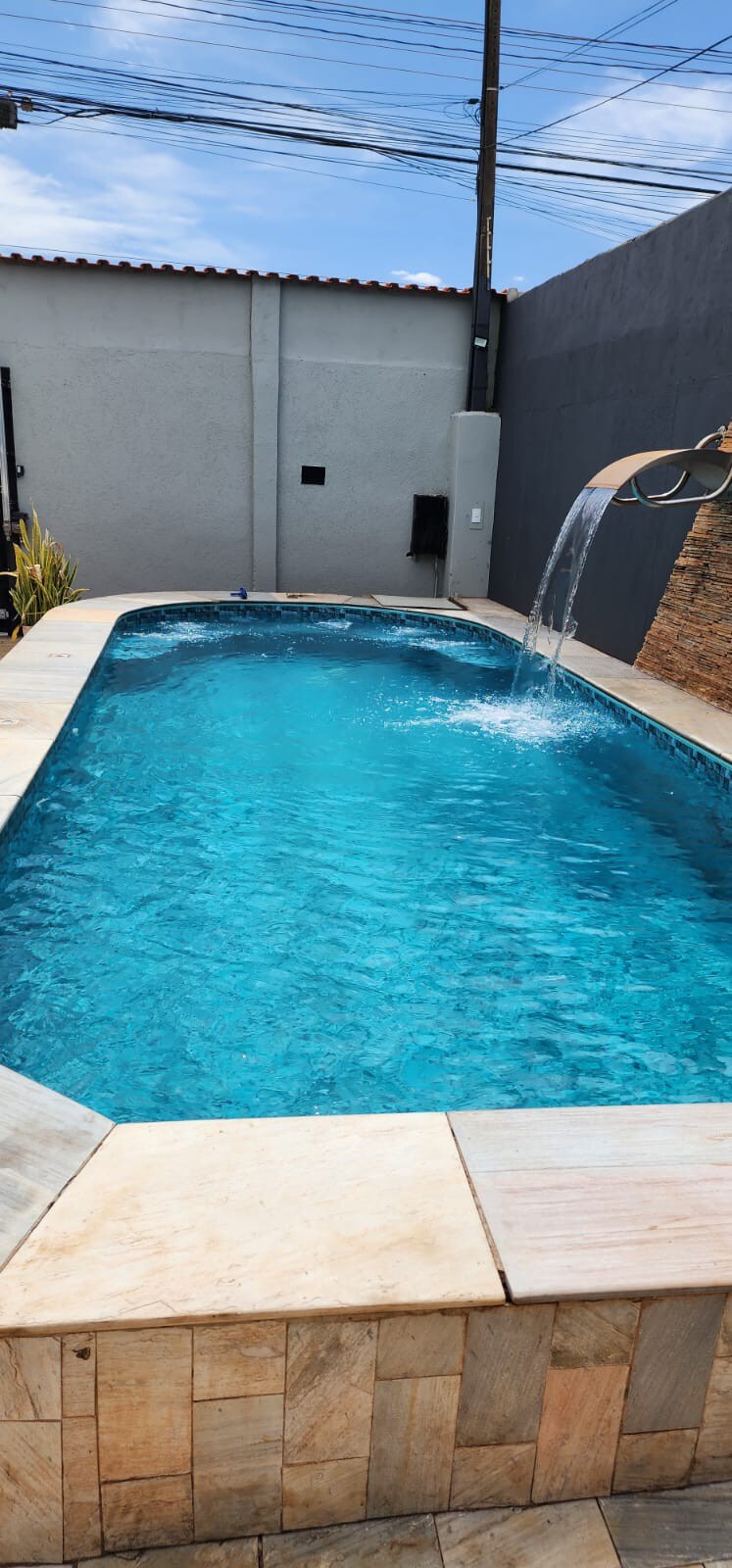 A spacious pool is featured, showcasing clear blue water and a gentle cascading waterfall. The pool area is bordered by textured stone tile, enhancing the relaxing environment. A wall in the background adds privacy, and sunlight reflects off the water's surface.