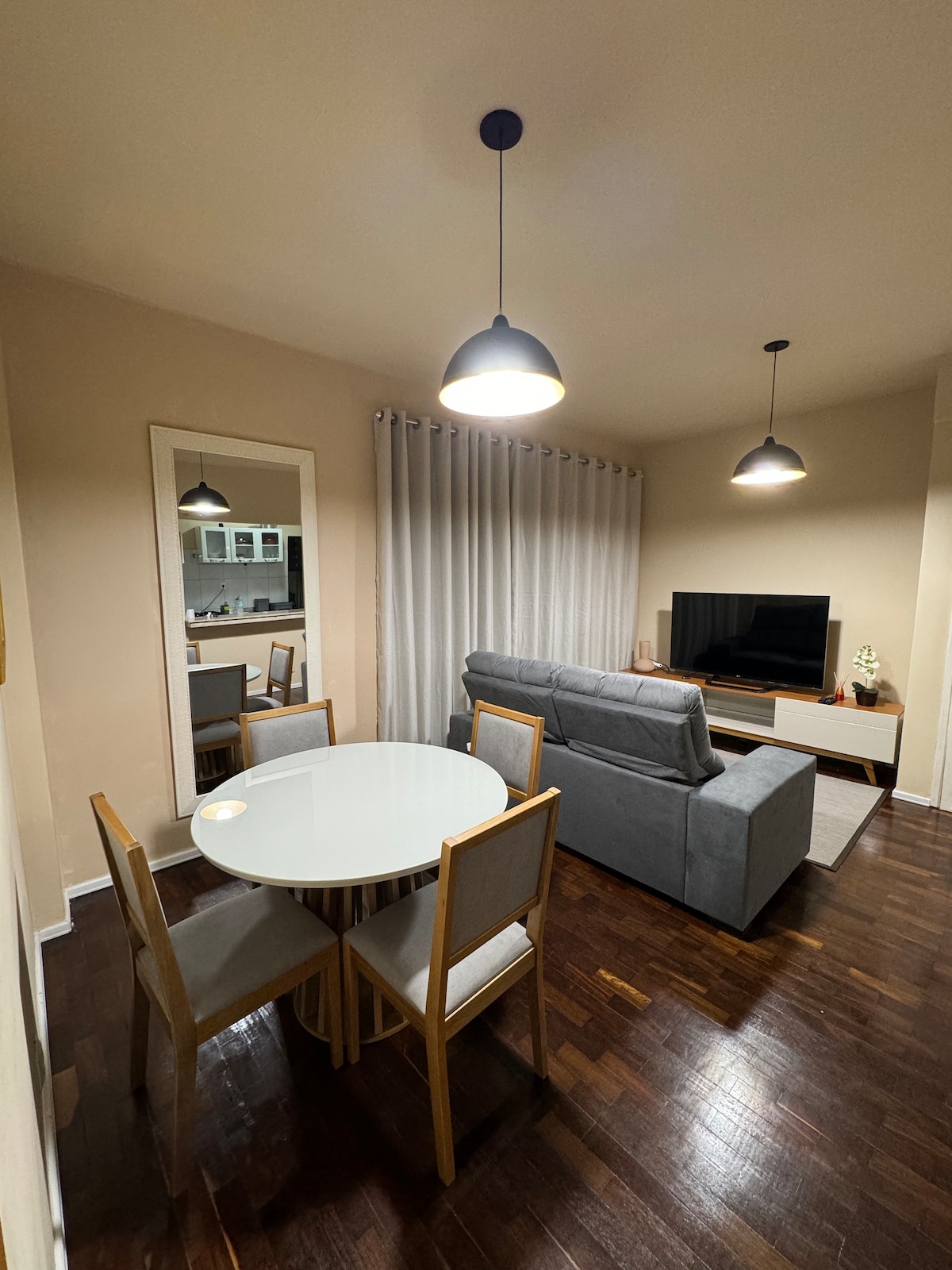 A dining area is depicted with a round white table surrounded by four light wooden chairs. A grey sofa faces a flat-screen TV, positioned against the wall. Soft lighting is provided by two pendant lights, creating a gentle glow in the space.