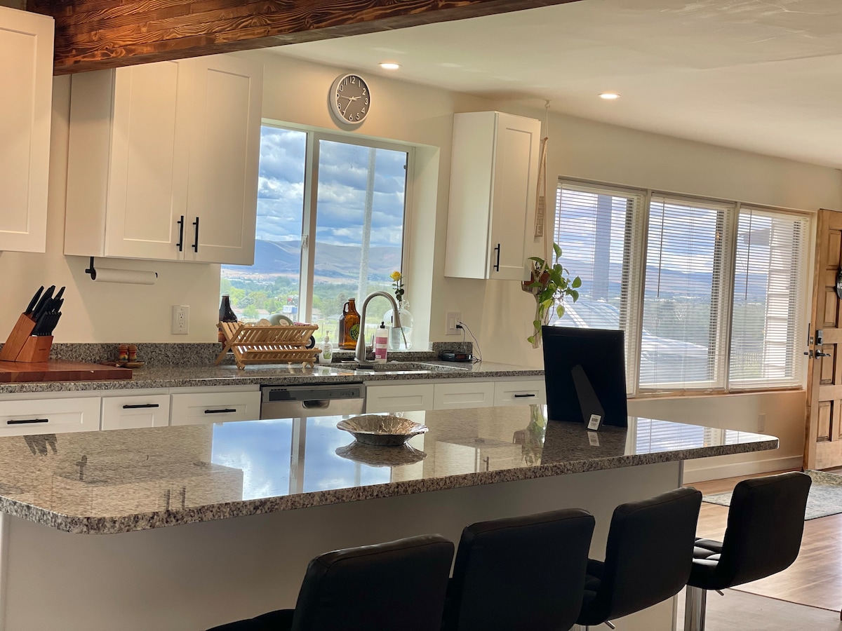 A spacious kitchen features an expansive granite island with seating. Light-colored cabinets and a modern design are complemented by a large window, providing a view of the outdoors. A sink and modern appliances are visible, enhancing functionality. Natural light floods the area through multiple windows.