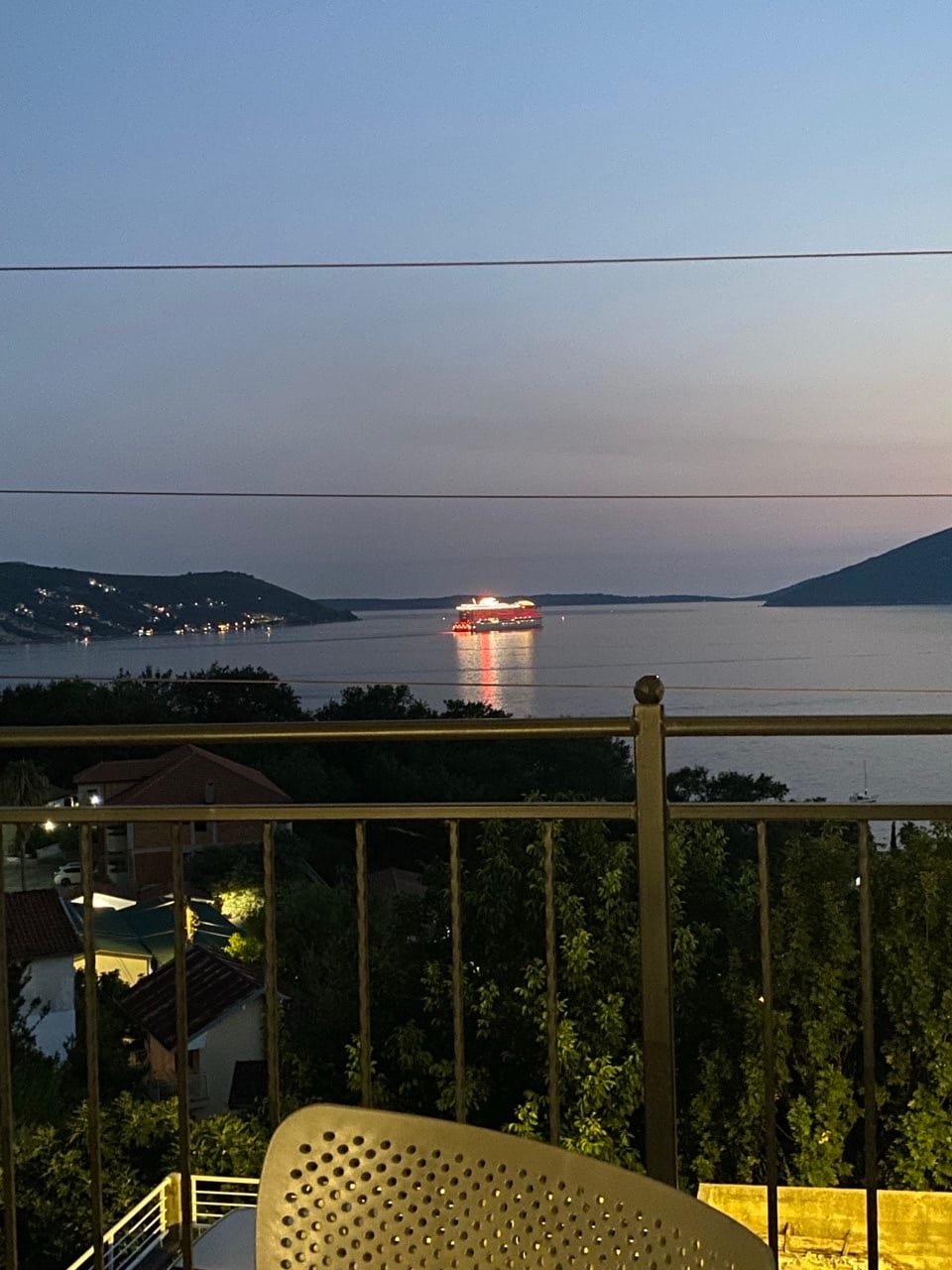 A panoramic view reveals a serene body of water reflecting soft twilight hues. Distant mountains outline the horizon, while a lit vessel gently glides across the surface. The scene is framed by the balcony railing, offering a peaceful glimpse of the surrounding landscape.