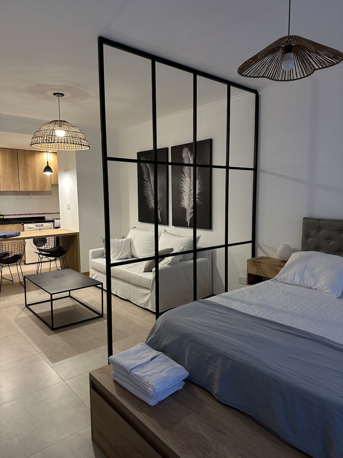 A modern sleeping area features a spacious bed with neutral linens, separated from a cozy living space by a stylish glass partition. A light-colored sofa and a coffee table contribute to the inviting atmosphere. Warm light fixtures provide illumination, enhancing the contemporary design.