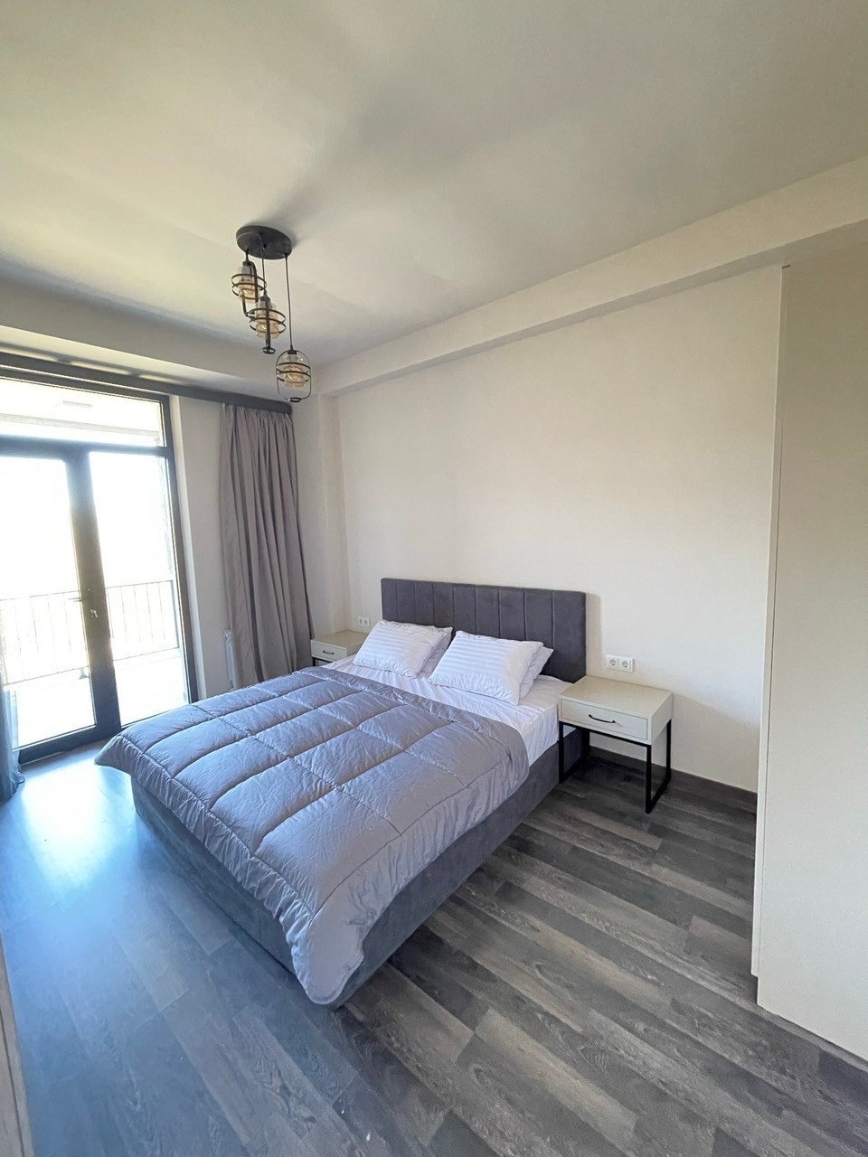 A serene bedroom is visible, featuring a gray upholstered bed with a neatly arranged comforter. Two bedside tables with lamps flank the bed. Large glass doors provide access to a balcony, with light filtering in through sheer curtains.