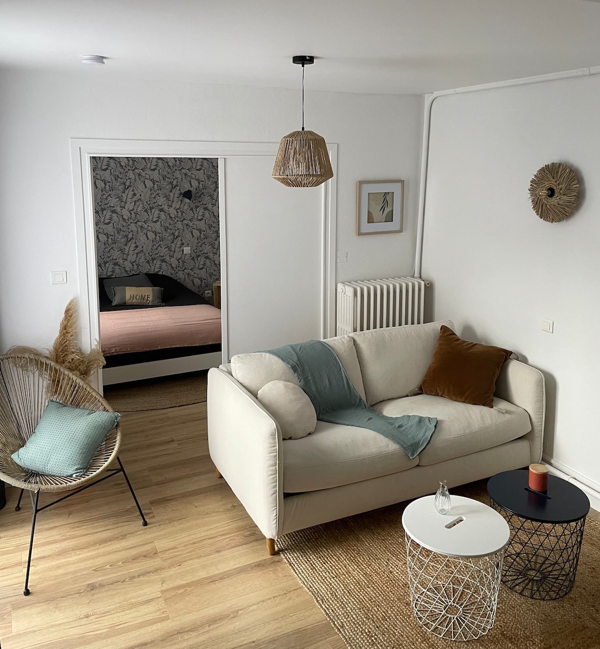 A well-lit living area features a white couch accompanied by a patterned throw and decorative cushions. A small, round black table supports a candle, while a basket-style side table adds texture. The background reveals a glimpse of a bedroom with a soft pink bed, framed artwork, and neutral décor.