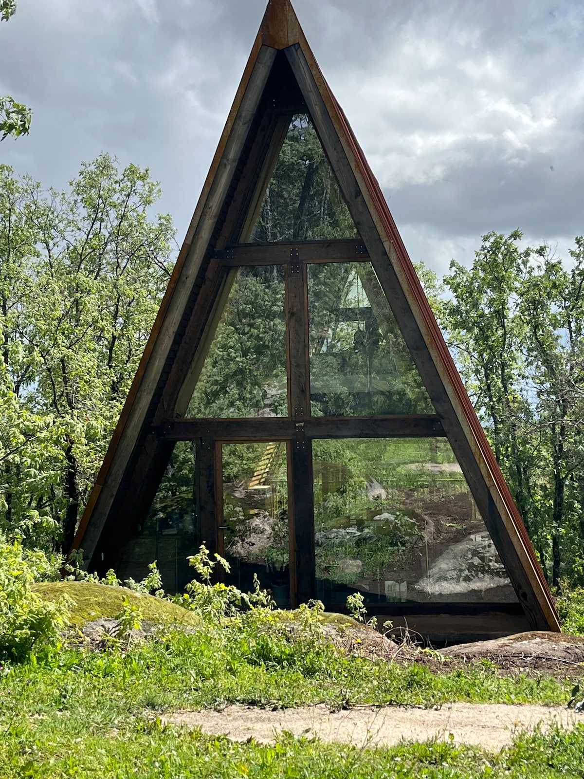 An A-frame cabin features large glass panels that reflect the surrounding greenery. Set against a backdrop of trees, the structure's peaked roof offers a unique architectural profile, while grass and wildflowers grow in the foreground.