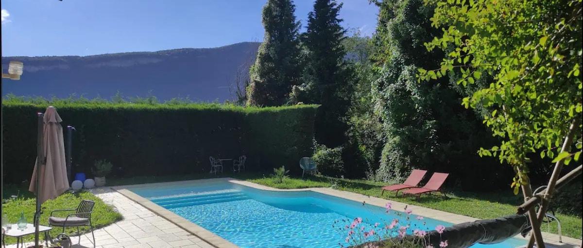 A private swimming pool is surrounded by a well-maintained garden, featuring lush greenery and neatly trimmed hedges. Two lounge chairs are positioned beside the water, with scenic views of mountains and trees in the background. A shaded seating area is visible, enhancing outdoor relaxation.