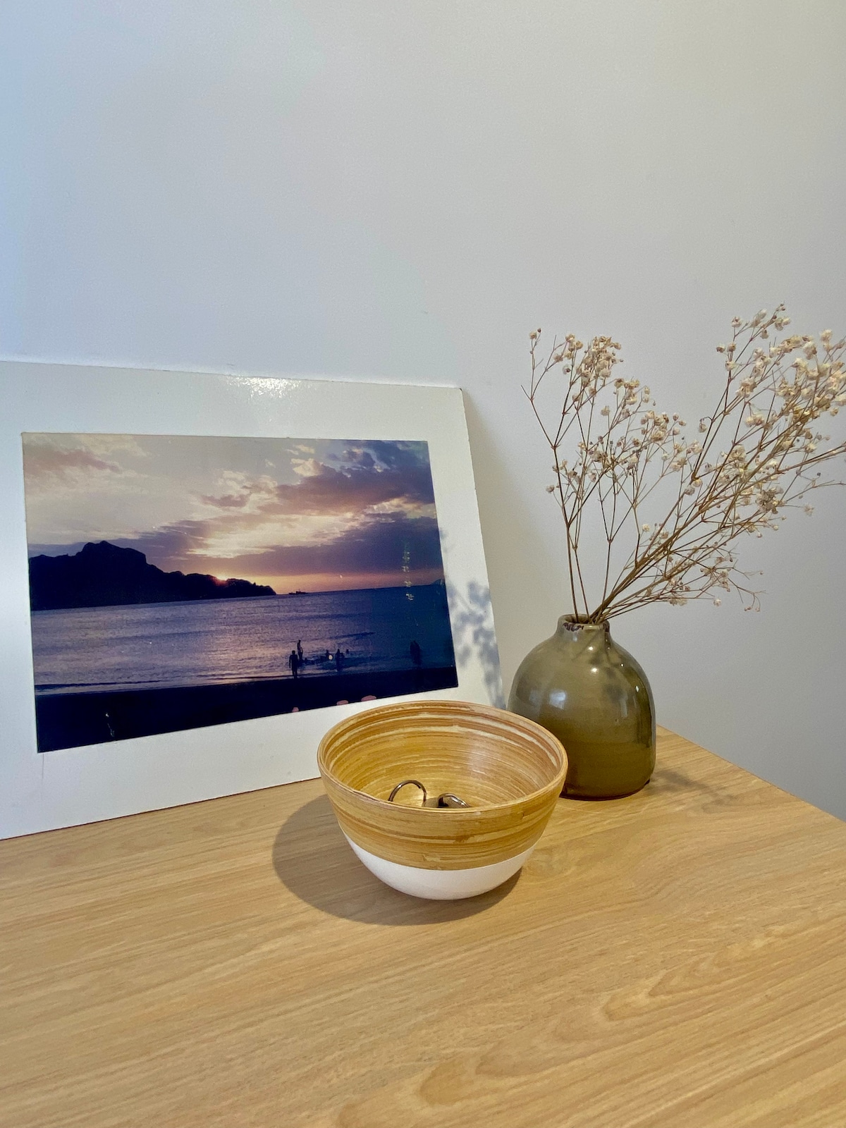 A warm wooden surface displays a bowl with a smooth, layered design. Next to it, a vase holds dried flowers, providing a subtle contrast. Behind, a framed photograph captures a serene sunset view over water, enhancing the overall aesthetic of the space.