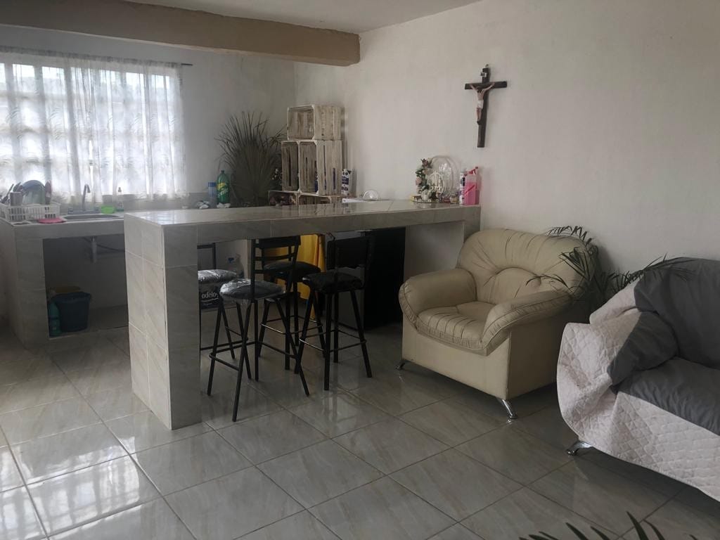 An open living space features a light-colored tiled floor and a large bar-like table with four high stools. A plush light sofa and a covered chair are situated nearby. Natural light filters through sheer curtains, illuminating the room's simple decor.