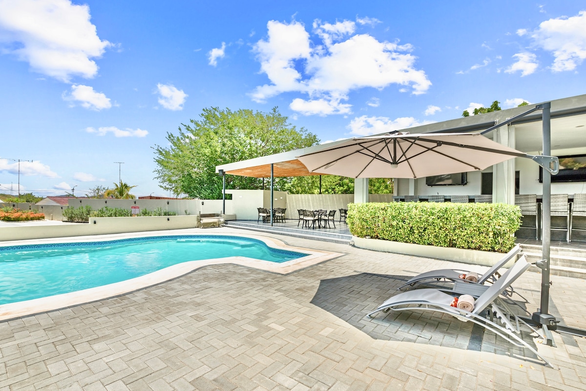 A spacious outdoor area features a private swimming pool surrounded by a tiled patio. Comfortable lounge chairs are positioned beside the pool, with an umbrella providing shade. Lush greenery and a dining table with chairs are situated beneath a canopy in the background.