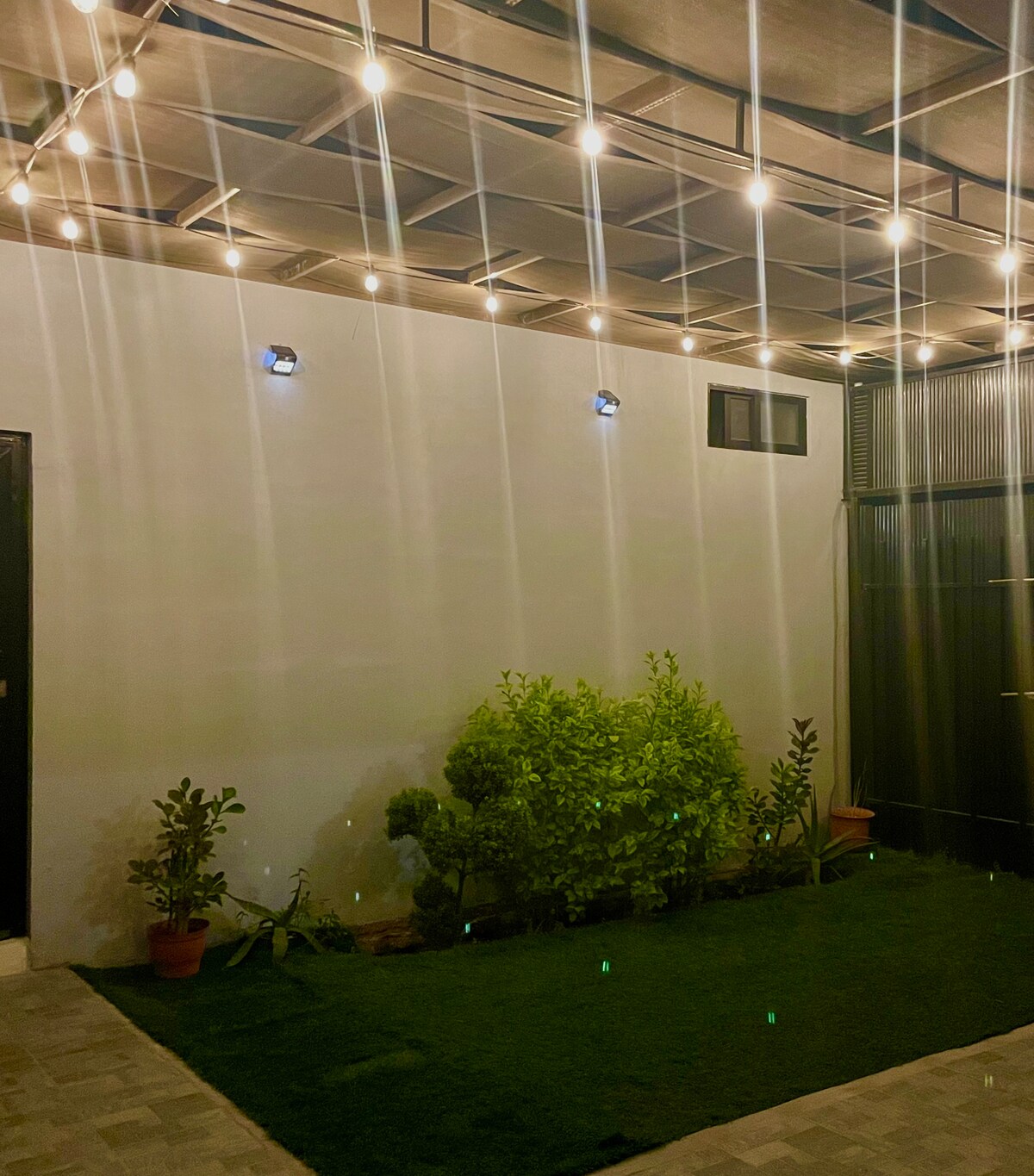A private outdoor patio area is illuminated by softly glowing string lights overhead. Low-maintenance plants are arranged in pots, adding greenery to the space. A wall with a small window is visible, enhancing the tranquil setting suitable for relaxation.