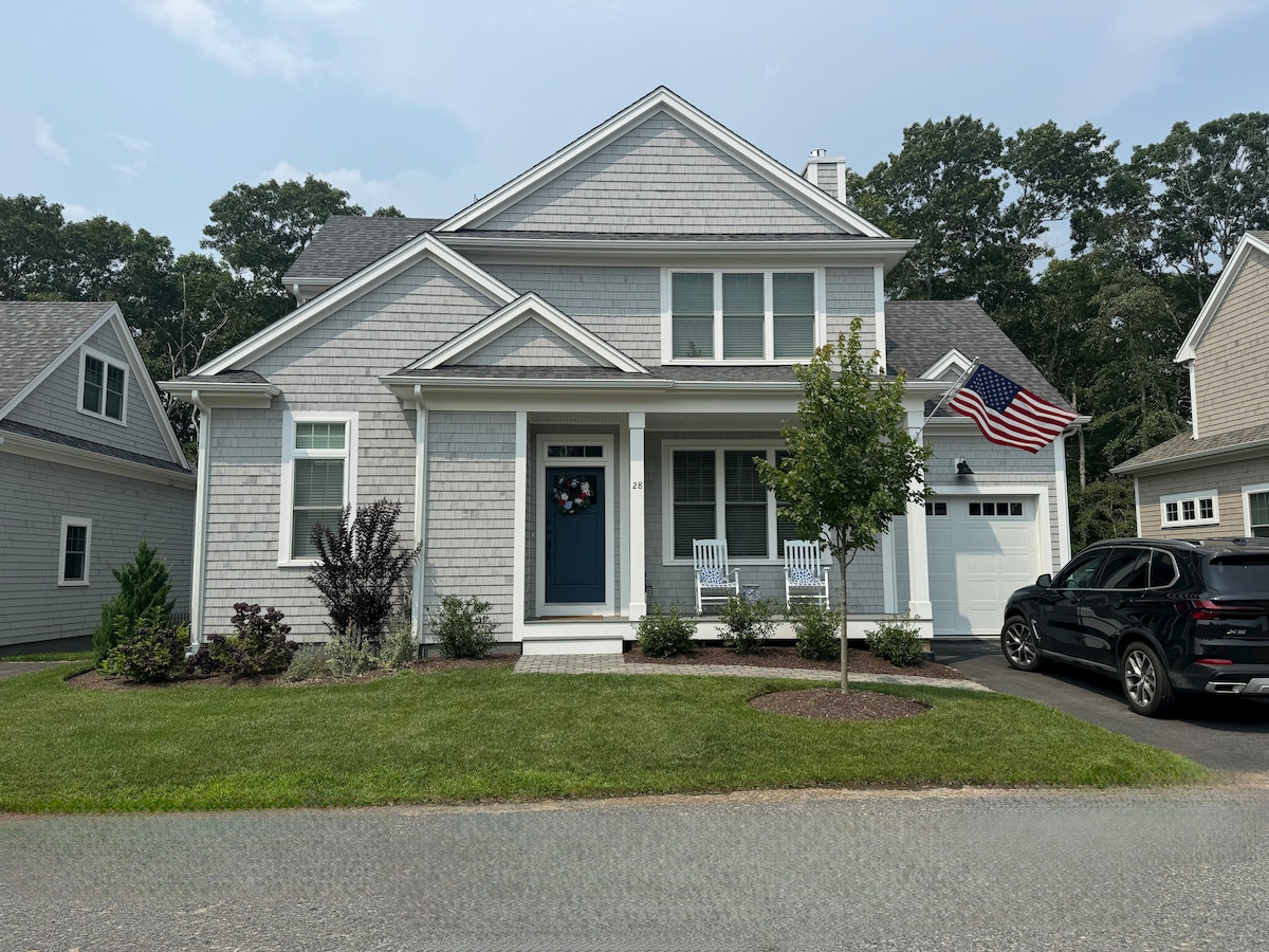 The Cape Cod-style home presents a light grey exterior with white trim and a welcoming front porch featuring two rocking chairs. A large American flag is displayed, and a neatly landscaped yard is visible, complemented by a paved driveway leading to an attached garage.