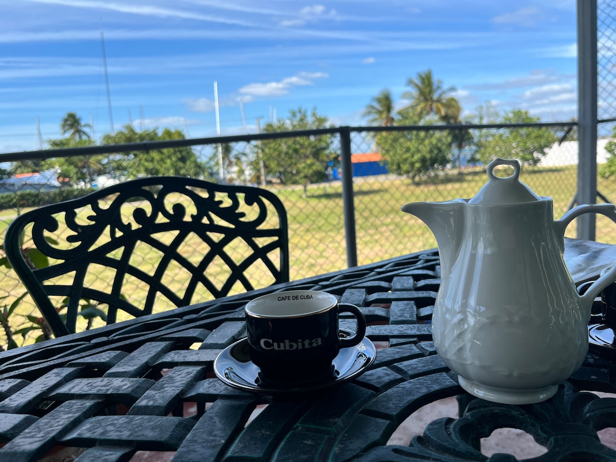 A decorative outdoor table features a black coffee cup labeled 'Cubita' alongside a white teapot and saucer. In the background, greenery and distant sailboats can be seen under a clear blue sky, creating a serene atmosphere for relaxation.