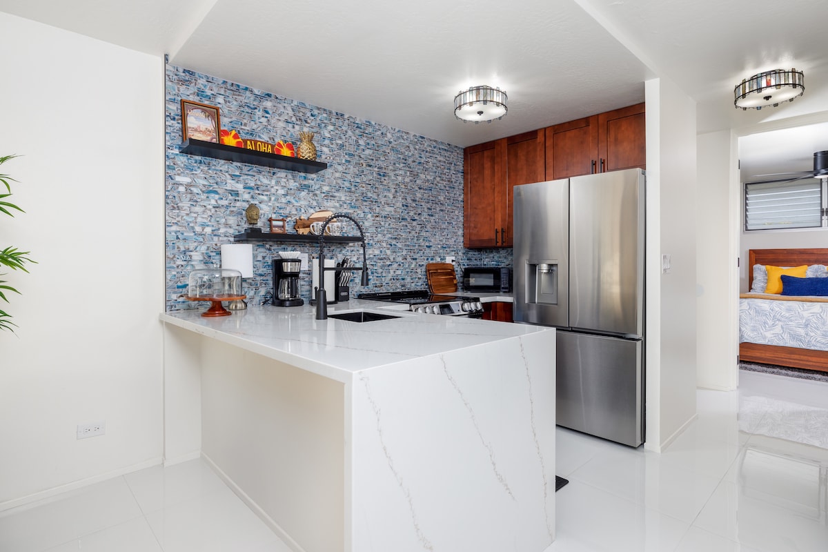 A modern kitchen features a spacious island with a white marble countertop, surrounded by wooden cabinetry. A decorative stone accent wall adds texture. New stainless steel appliances, including a refrigerator and stove, are visible, alongside coffee-making equipment on the counter.