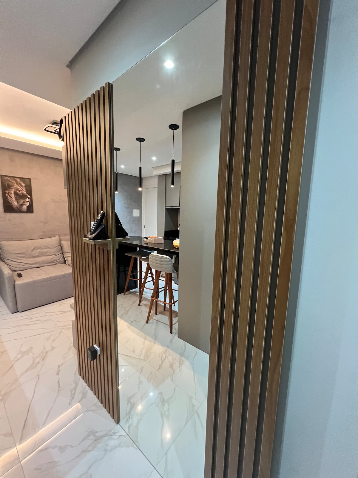 A modern entryway is showcased, featuring a wooden door with vertical slats. The space leads into a bright room with sleek furniture, including a light-colored sofa and dining area with high chairs, set against a backdrop of subtle lighting and polished tiled flooring.