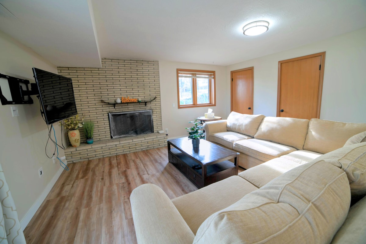 A comfortable living area features a large sectional couch and a coffee table, providing a cozy space for relaxation. A flat-screen TV is mounted on the wall opposite a brick fireplace, complemented by natural light from the nearby window.