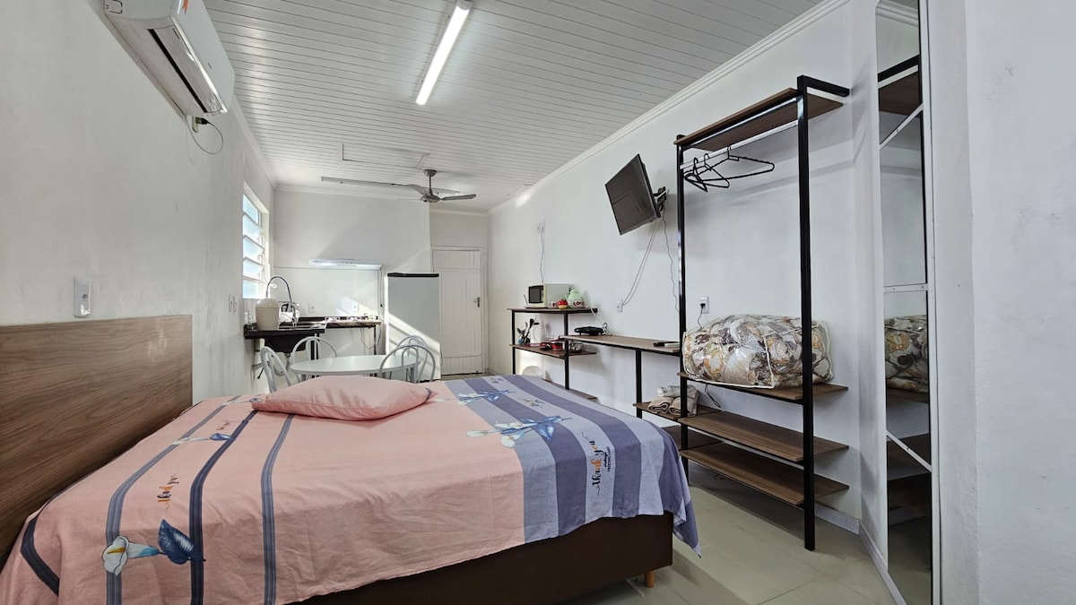 A spacious sleeping area features a large bed with a soft pink and blue striped blanket. Nearby, a television is mounted on the wall, and a clothing rack provides storage. A well-lit kitchen space is visible in the background, complemented by dining chairs and a table.
