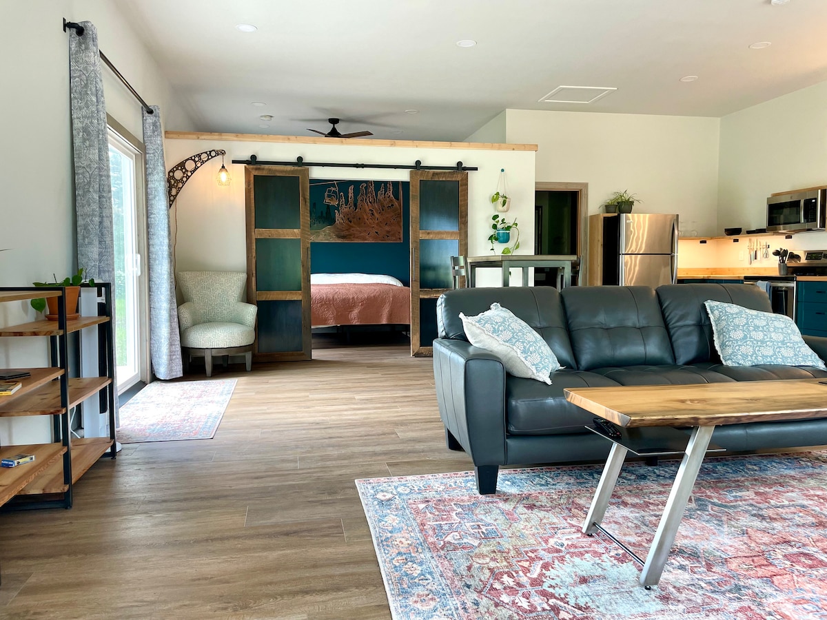 An open living space features a comfortable sofa with decorative pillows, a stylish coffee table, and a vibrant area rug. Large sliding glass doors provide natural light, leading to outdoor views. A cozy sleeping area is partially visible in the background, separated by sliding panels.