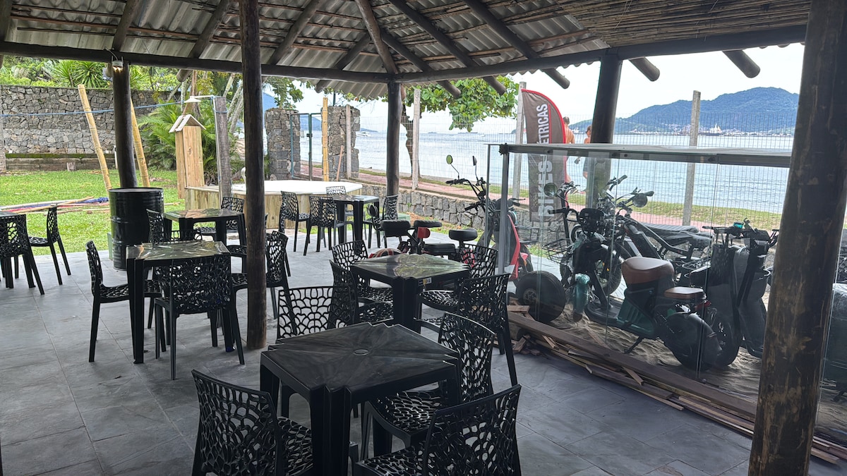 An outdoor seating area is covered by a rustic roof, featuring several black tables and chairs arranged for dining. A view of the beach and distant hills is visible beyond the railing. Bicycles and scooters are parked nearby, offering convenient mobility options.