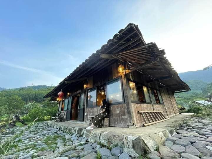 A rustic wooden house is situated on a stone pathway, surrounded by lush greenery and mountains. Large windows allow for views of the landscape. Lanterns hang outside, adding a touch of character to the structure, which exudes a tranquil and natural ambiance.