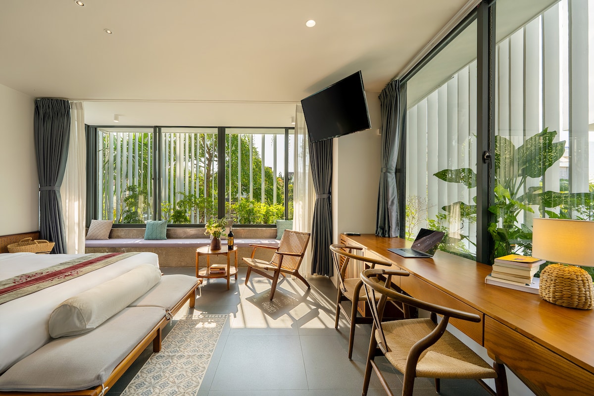 A well-lit room is showcased with large windows allowing views of lush greenery. A spacious bed is positioned beside a comfortable seating area, complemented by two wooden chairs. A sleek desk with a laptop and decorative items is also present, enhancing the room's modern design.