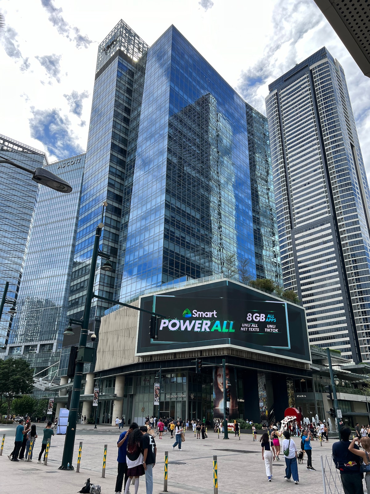 A modern urban landscape is captured, showcasing a cluster of tall glass buildings reflecting clouds in the sky. The structures display a sleek design, with vibrant advertisements adorning the lower levels. People are seen walking in the vicinity, highlighting the bustling atmosphere.