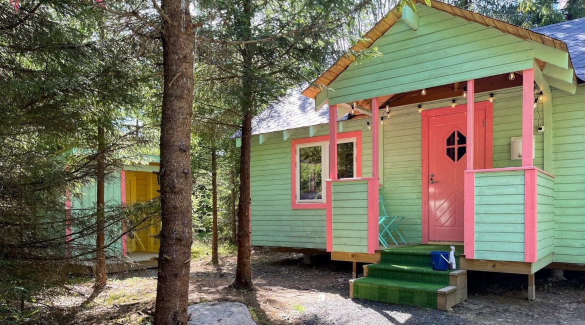 A charming green cabin is framed by tall trees, featuring pink accents around the door and windows. A small porch with a green mat invites entrance, while an adjacent colorful structure adds to the vibrant exterior. Natural surroundings create a tranquil setting.