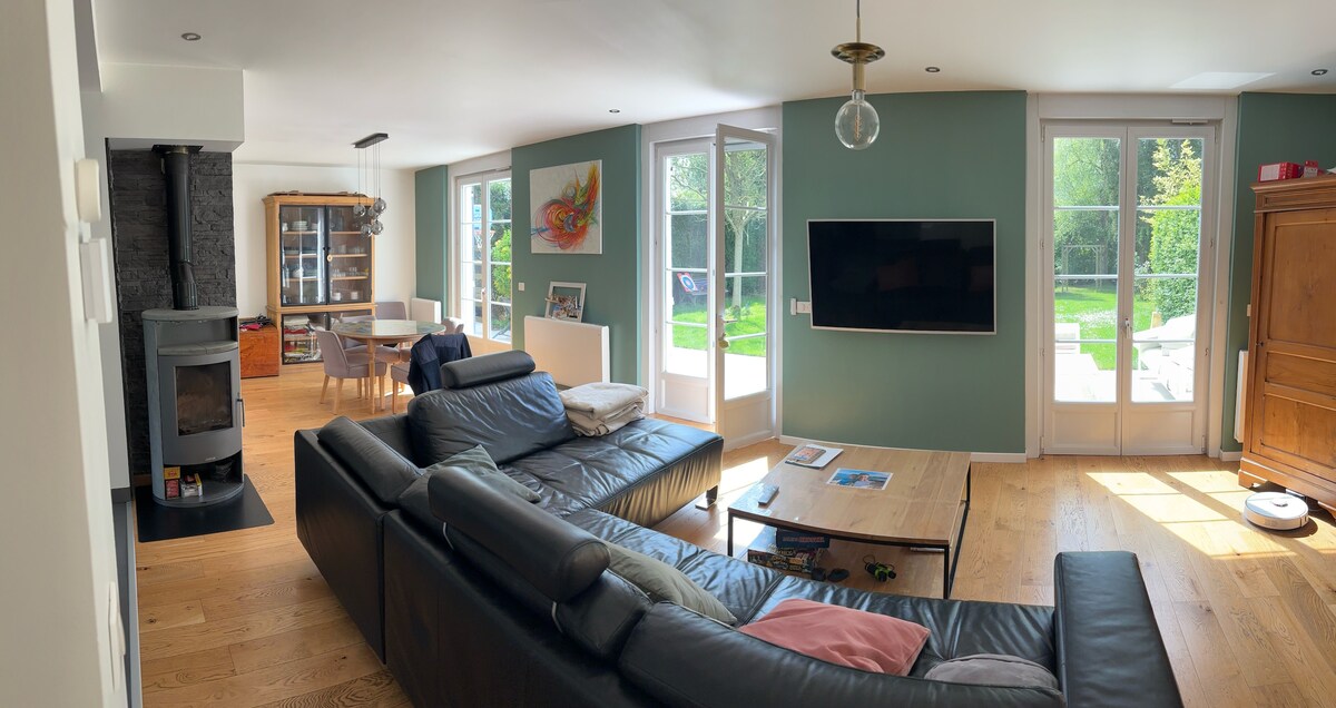 A modern living area features a large sectional sofa arranged around a wooden coffee table. Natural light fills the space through multiple glass doors, leading to a garden. A wall-mounted television and a cozy wood stove enhance the welcoming atmosphere.