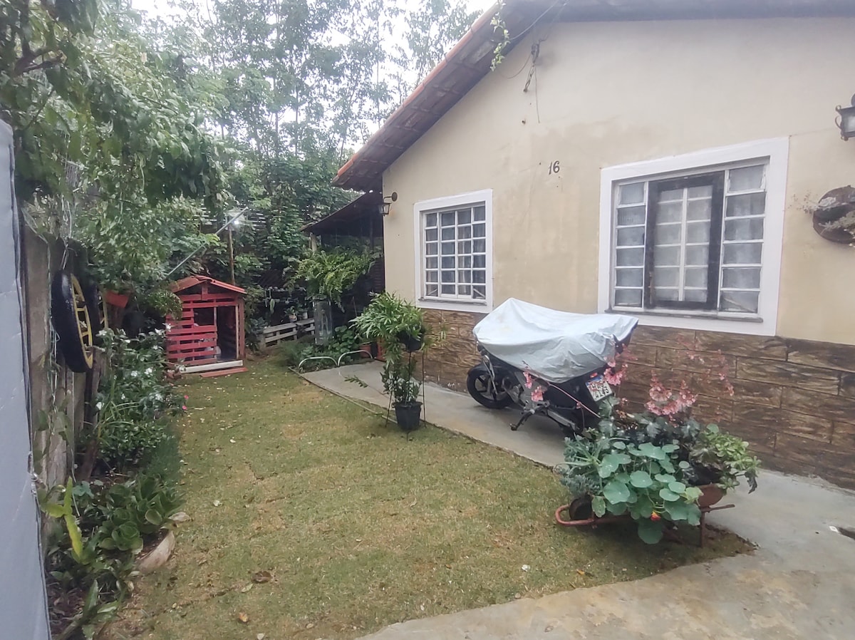 A peaceful outdoor area showcases a well-maintained lawn surrounded by lush plants. The side of the house is visible, featuring two windows adorned with white grilles. A covered motorcycle is parked on a concrete path, while a small playhouse can be seen in the background.