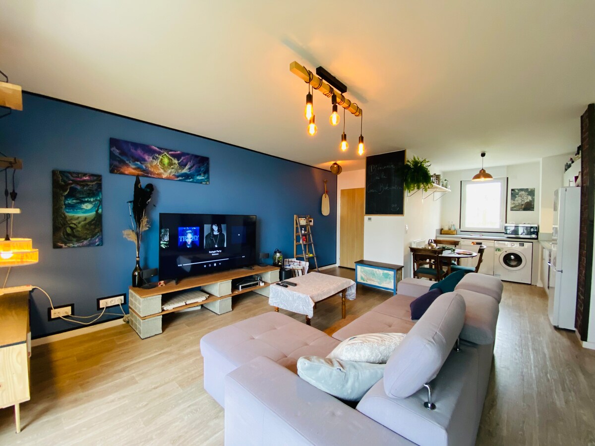 A spacious living area is featured, highlighted by a large screen TV and modern light fixtures. The decor includes artistic wall paintings against a blue accent wall. A comfortable sofa with cushions invites relaxation, and the adjoining kitchen area is visible in the background.