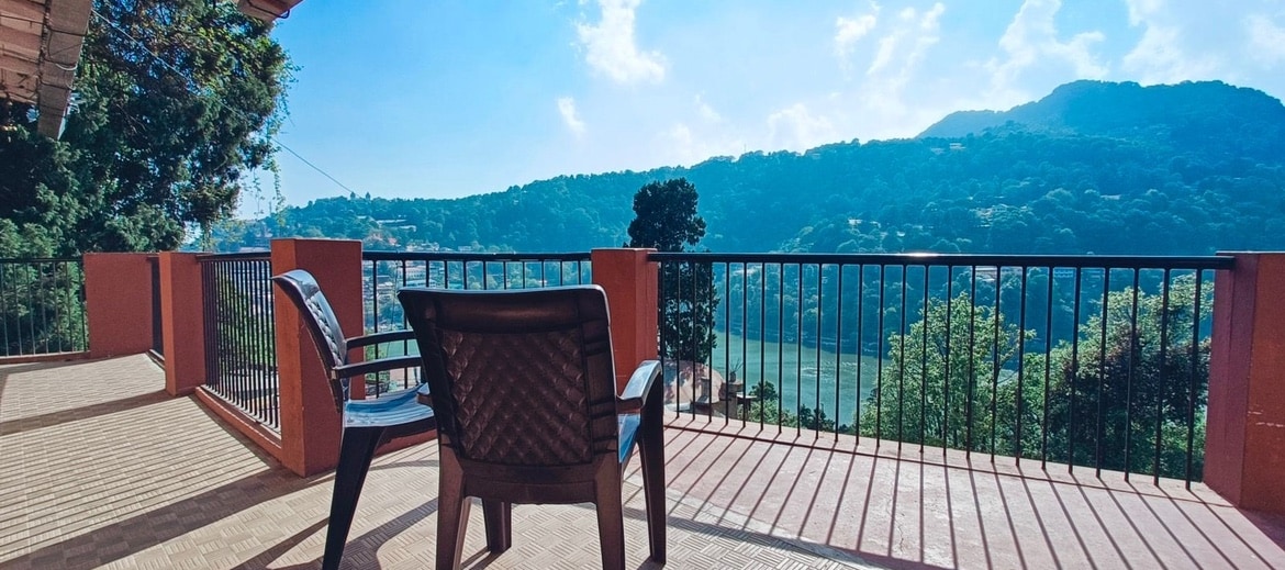 A spacious balcony features a single chair positioned to take in expansive views of Naini Lake and the surrounding hills. The floor is covered with a textured surface, and a railing offers safety while enjoying the serene landscape.