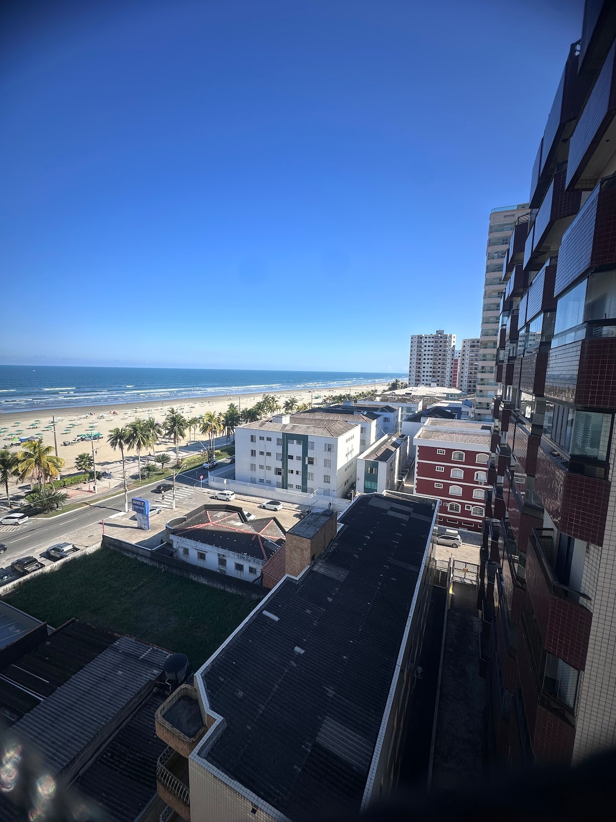An expansive view showcasing the beach and ocean, framed by blue sky. A coastal skyline is visible, featuring palm trees lining the sandy shore. Various buildings are positioned along the street, with a mix of residential and commercial structures in the foreground.