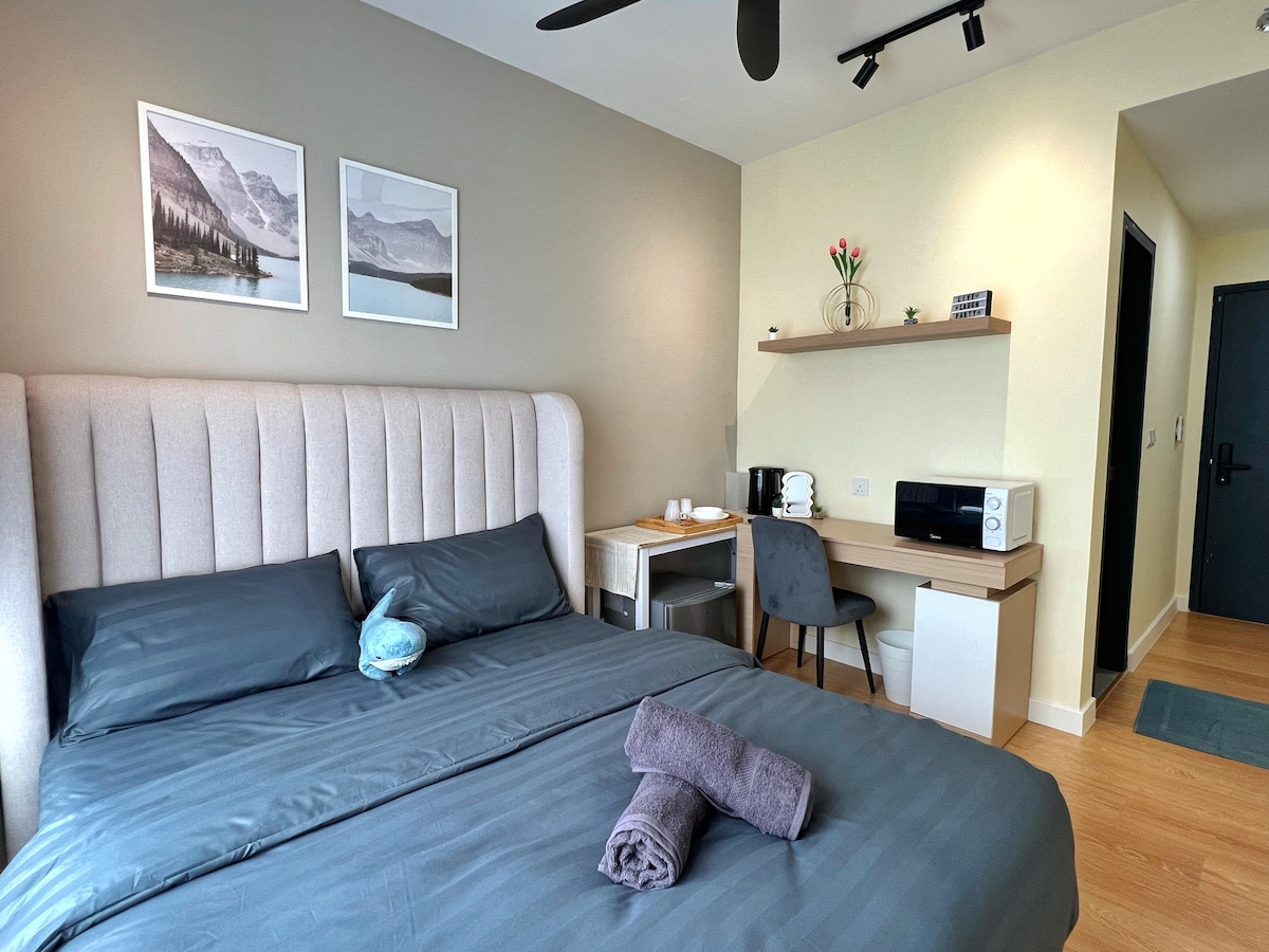 A serene bedroom features a comfortable queen-sized bed with soft linens and neatly arranged towels. A cozy table and chair set is positioned beside the kitchenette, which includes a microwave. Two framed mountain-themed artworks are displayed above the headboard, adding a touch of nature.