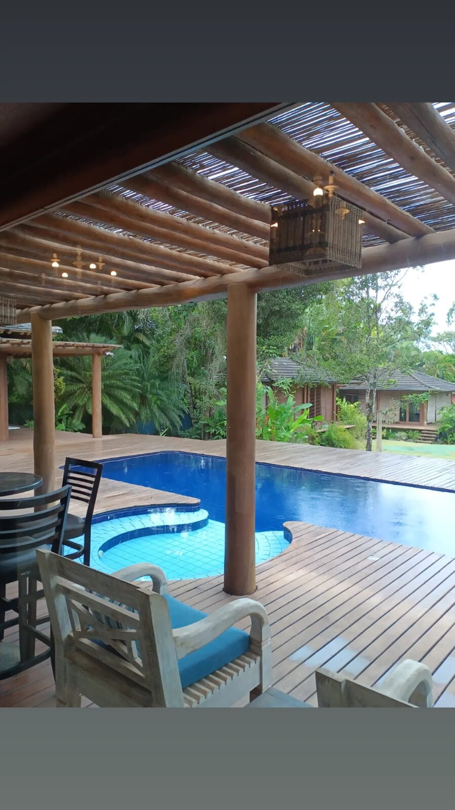 A covered terrace showcases a sparkling blue pool surrounded by wooden decking. On one side, a dining area features a table with multiple black chairs. Lush greenery and additional structures are visible in the background, adding to the tropical setting.