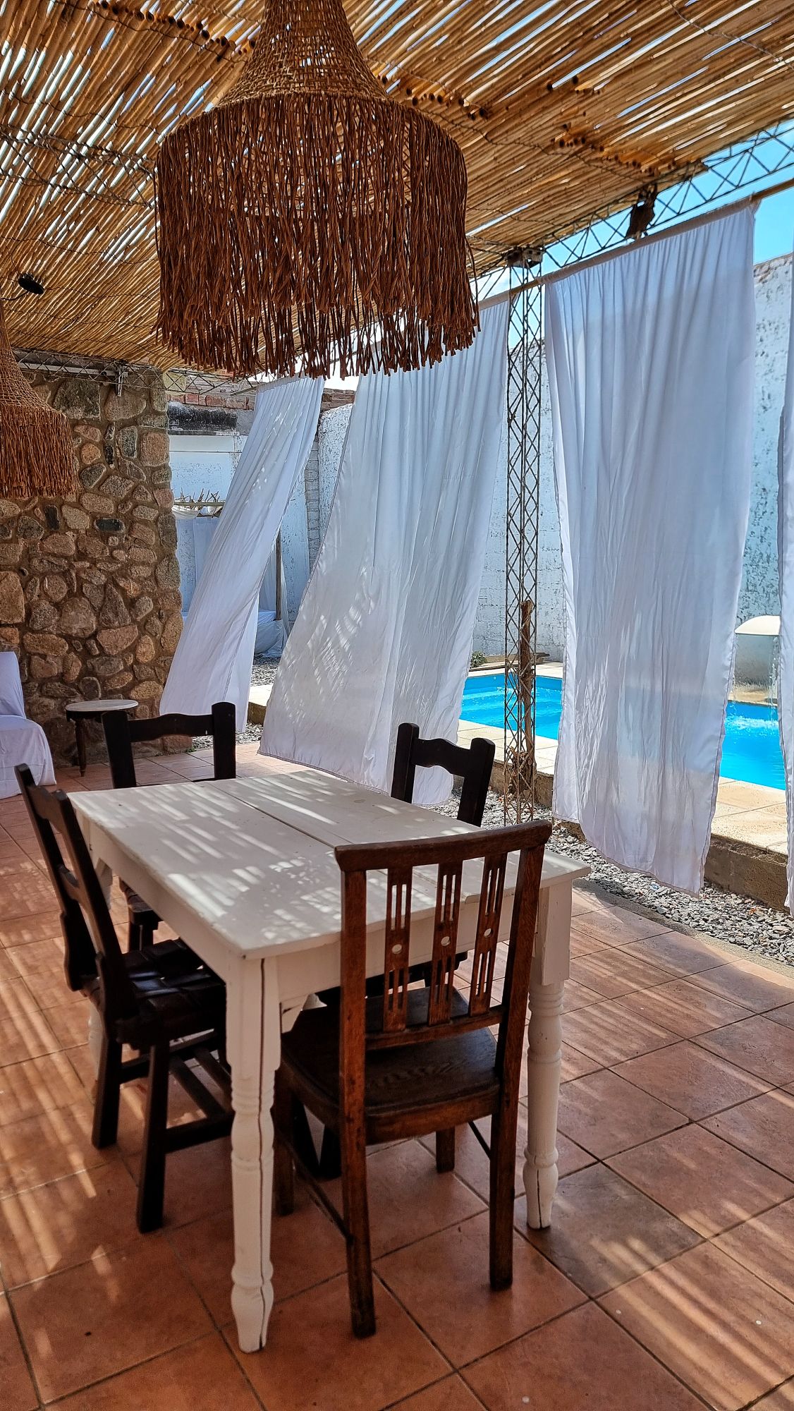 An outdoor dining area features a white table surrounded by wooden chairs. Sunlight filters through a straw canopy, casting patterned shadows on the tiled floor. White curtains flutter gently in the breeze, revealing a glimpse of a tranquil blue pool nearby.