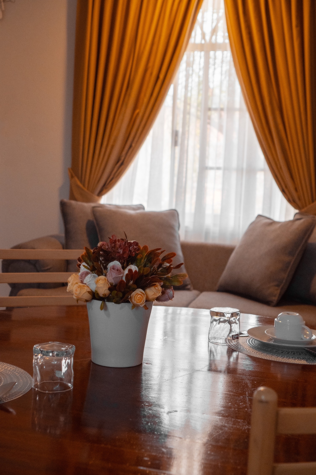 A dining table is set with clear glassware and white dishware, surrounded by neutral-colored chairs. A vase filled with fresh flowers adds a pop of color, while warm curtains frame a bright window casting soft light onto the polished wood surface.