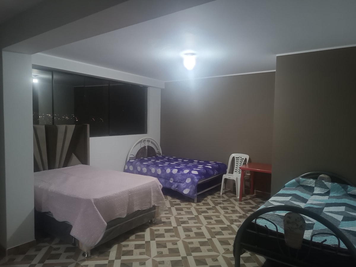 A spacious bedroom is featured, containing three beds with colorful bedding. A table and a white plastic chair are positioned against the wall. Large windows allow natural light, offering a view of the surroundings. The floor is decorated with geometric tile patterns.