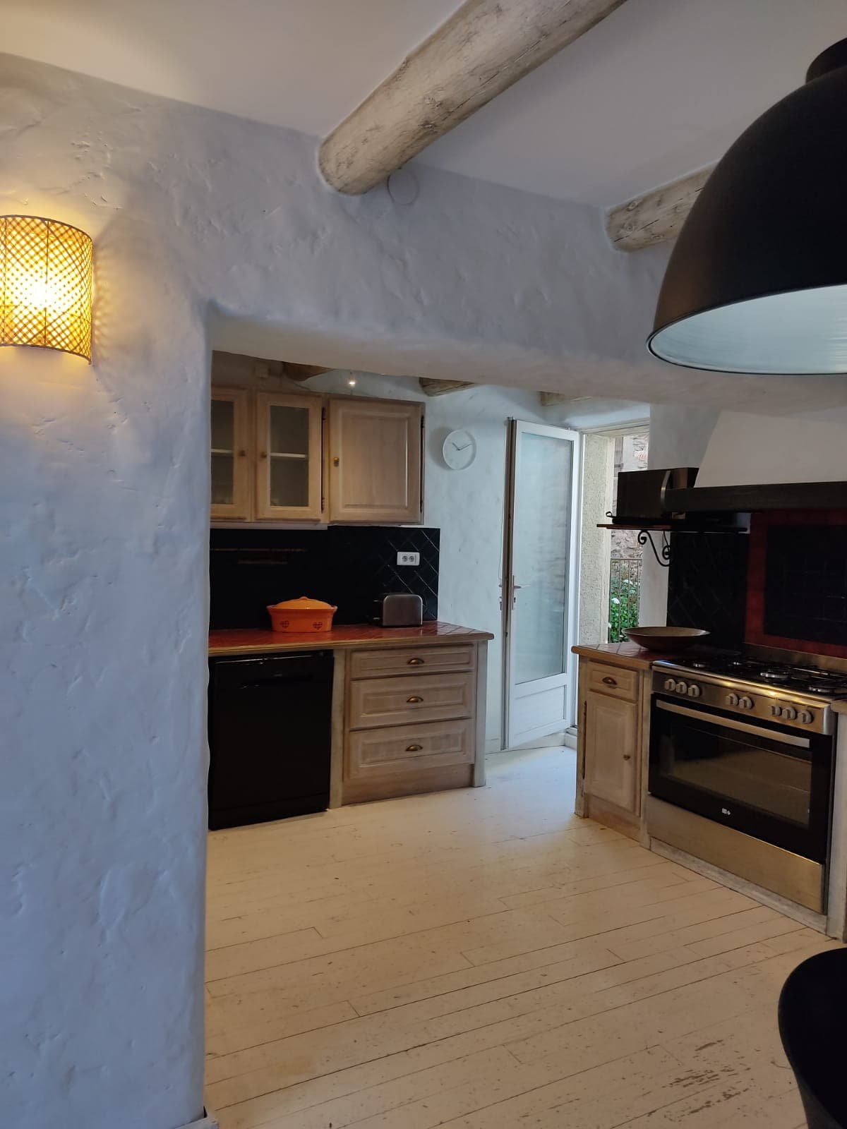 A functional kitchen space features light-colored wood cabinetry with a mix of modern and rustic design elements. Large appliances, including an oven and refrigerator, are present. Natural light enters through an adjoining doorway, creating a warm ambiance. A pendant light hangs overhead, adding a cozy touch.