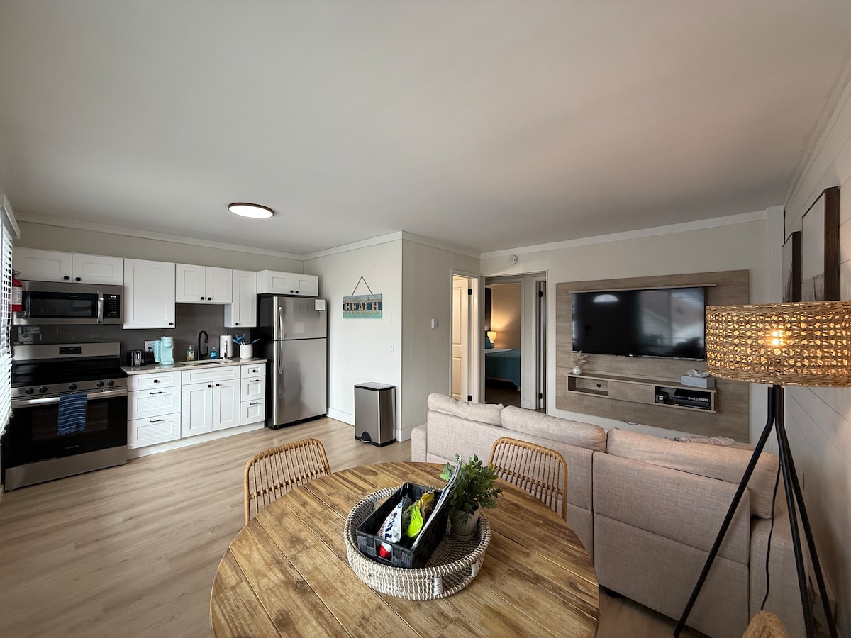 An open living space is showcased, featuring a modern kitchen with stainless steel appliances and white cabinetry. A comfortable sofa faces a wall-mounted television. A round dining table with woven chairs sits in the foreground, adorned with a decorative tray holding various items.