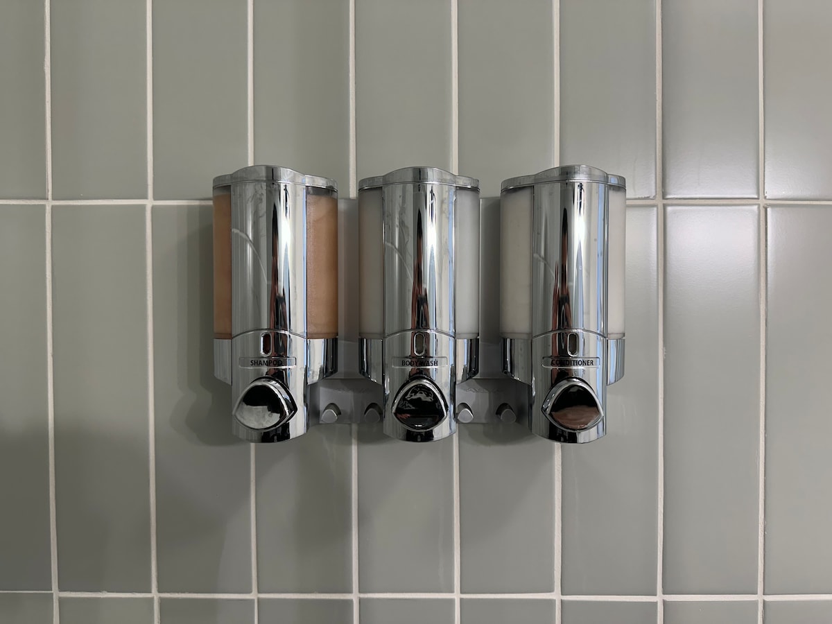 Three chrome shower dispensers are mounted on a tiled wall with light green tiles. Each dispenser is filled with liquid soap, shampoo, and conditioner, providing guests with convenient access to bathing essentials.