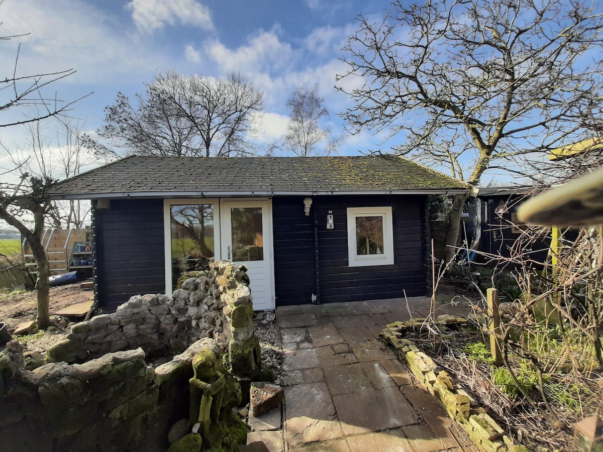A charming exterior of a wooden house is displayed, featuring a dark facade and a sloped roof. A small entrance is visible, framed by a stone pathway leading to the door. Nearby, trees and greenery surround the property, adding to the tranquil setting.