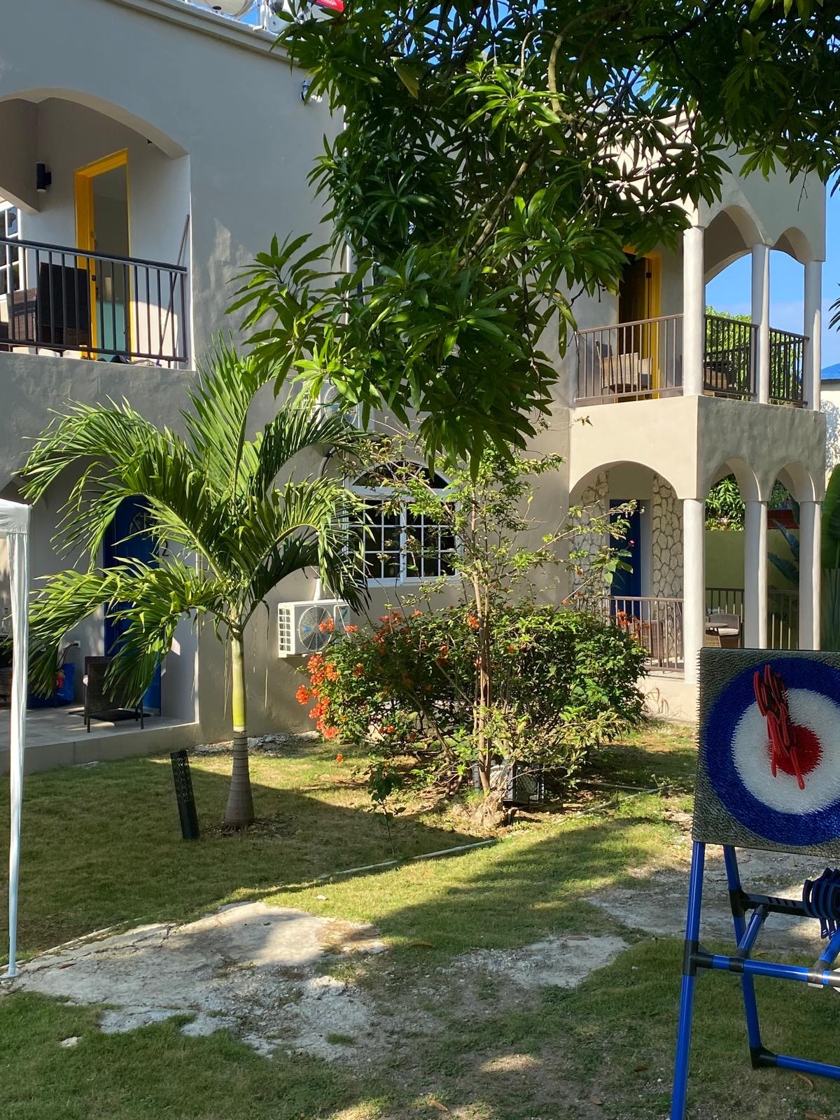 The property showcases a two-story structure surrounded by well-maintained gardens. Tropical plants and colorful flowers add to the landscape, while a palm tree stands prominently. The balcony displays vibrant blue and yellow doors, inviting guests to explore the charming outdoor space.