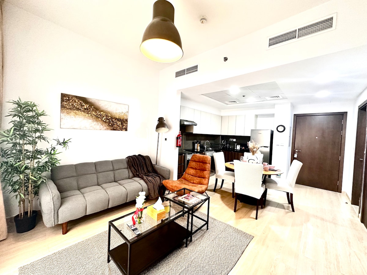 A modern living area features a gray sofa and an orange armchair beside a glass-top coffee table. A dining table is set for four, located in the adjacent open kitchen. Bright natural light fills the space, and decorative plants add warmth.