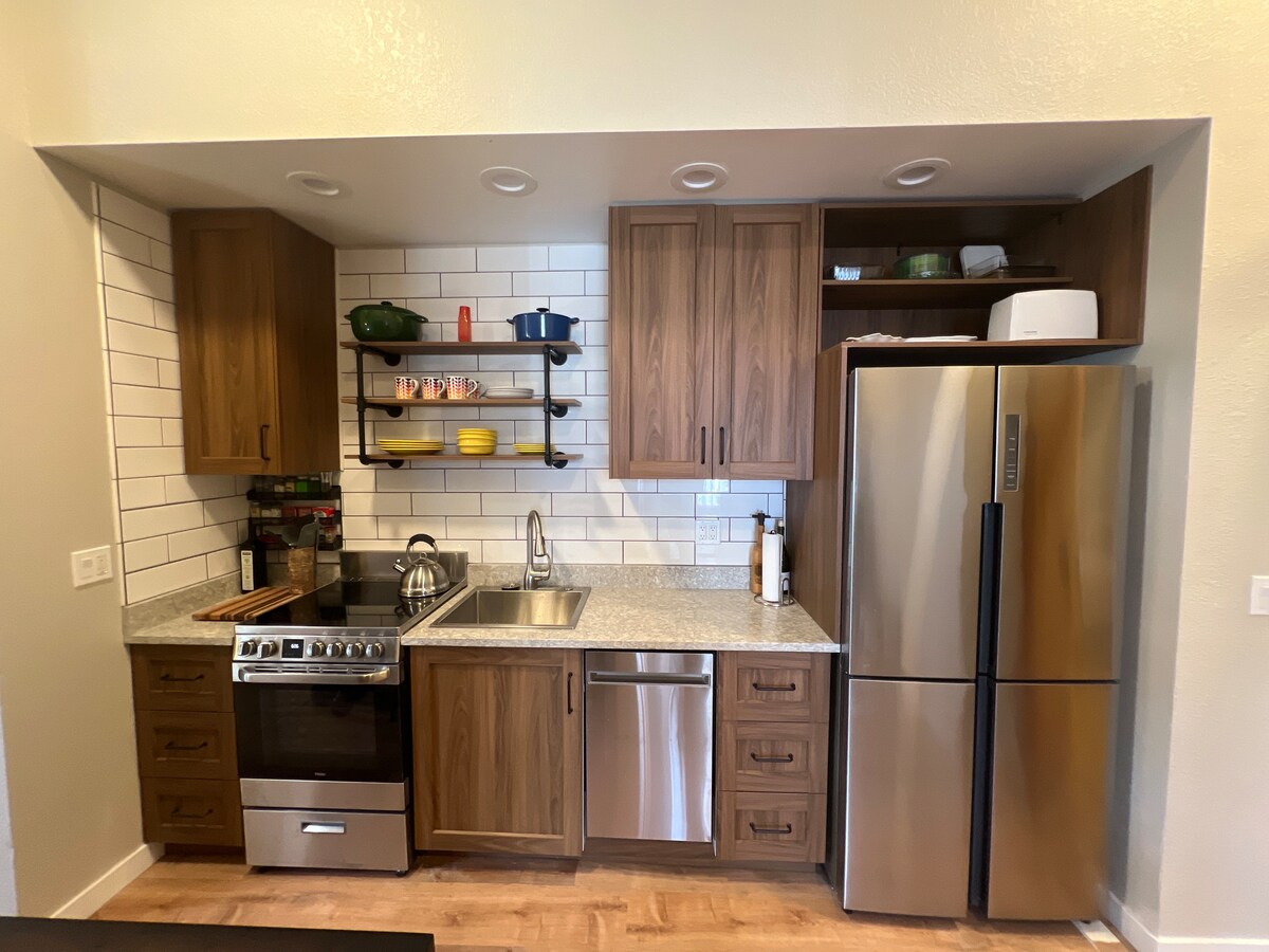 The kitchen features modern wooden cabinetry complemented by a stainless steel refrigerator and oven. A sleek countertop provides space for meal preparation. Open shelving displays colorful dishes, and a well-organized layout enhances functionality.